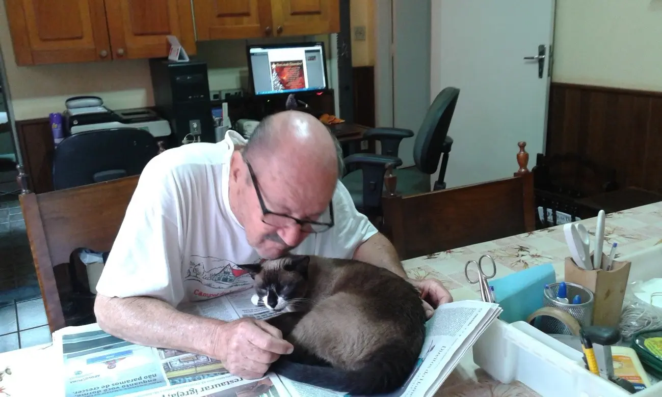 Eurípedes Kuhl com a gata Pituca. Descrição: Eurípedes sentado na mesa com jornal aberto na frente dele. A gata siamesa está deitada em cima do jornal e recebe afagos do escritor. Foto: arquivo pessoal