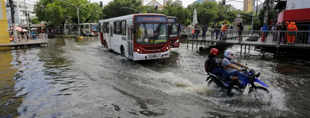 Rua alagada, ônibus e motos circulando.