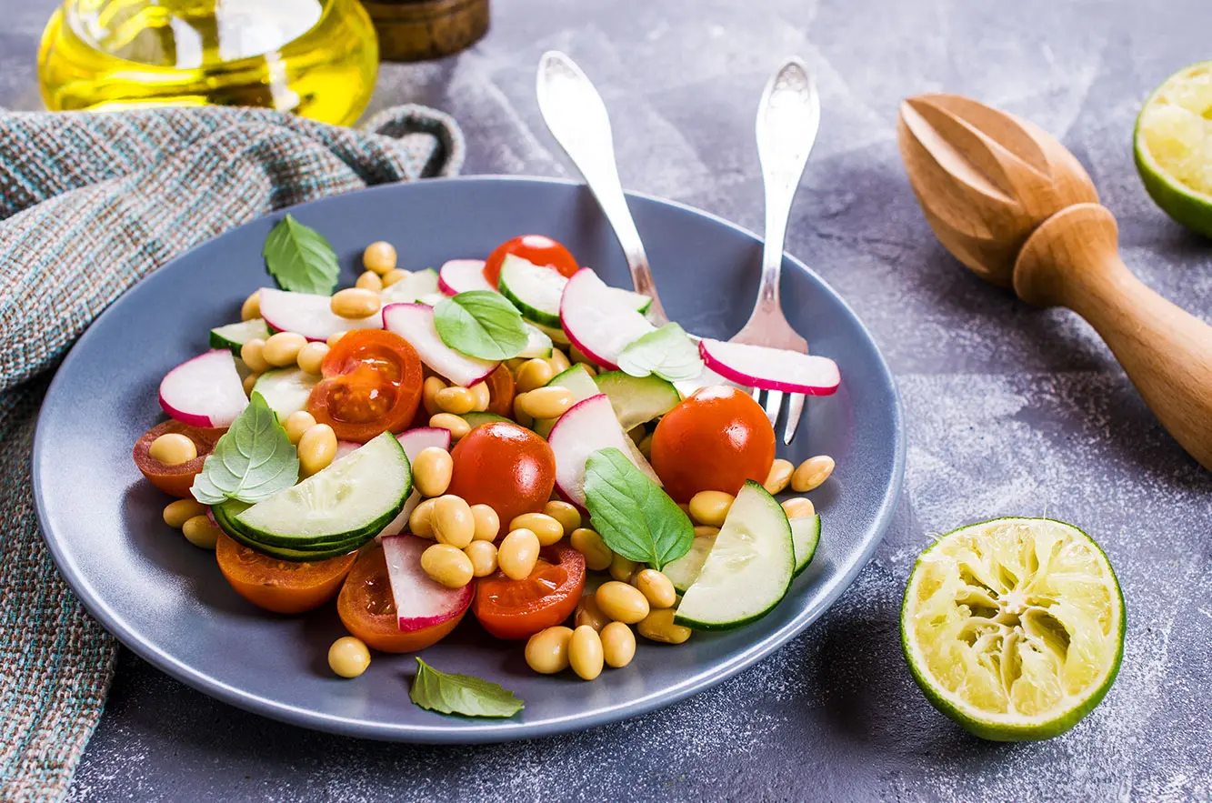 Prato cinza com soja, mini tomates, pepino e folha de manjericão; dentro do prato há dois talheres. Compondo o cenário há um limão sem o suco, um pano de prato cinza, azeite em um fracos de vidro redondo e um enfeite . 