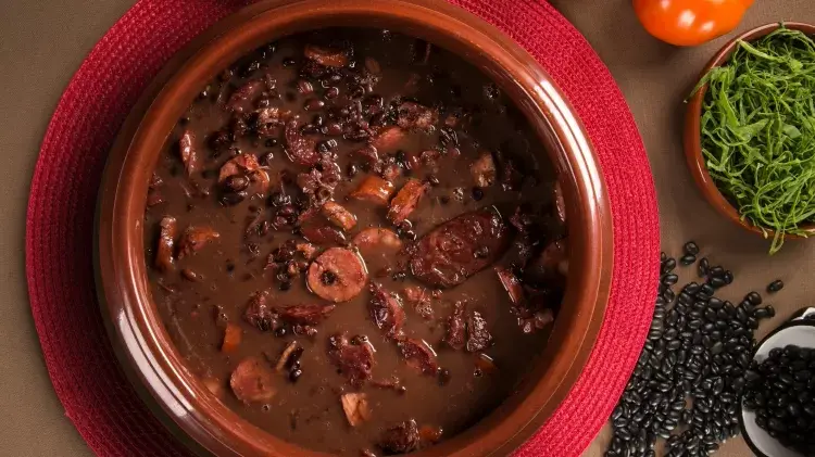 Prato marrom com feijoada em cima de um jogo americano vermelho.