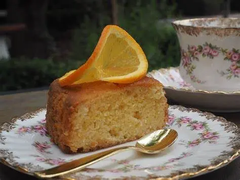 Bolo de laranja com uma rodela de laranja no topo, em um pratinho decorado com folhes rosas e uma colher ao lado.