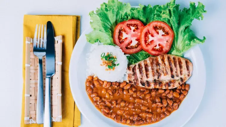 Prato com arroz, alface, duas rodelas de tomate, frango grelhado e feijão, ao lado um guardanapo amarelo e em cima garfo e faca.