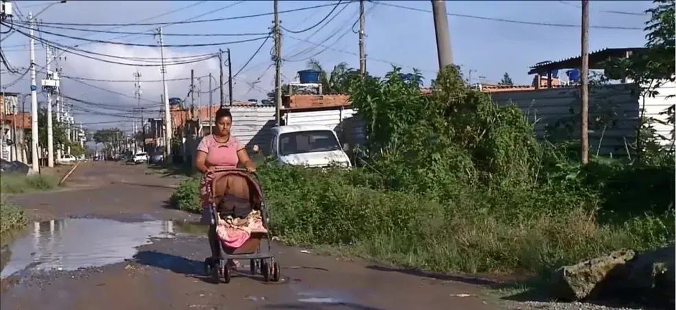 A imagem mostra uma moça, cabelo preso e vestindo roupa rosa, empurrando um carrinho de bebê. Atrás dela há um pouco de ver, postes de luz e alguns carros estacionados.